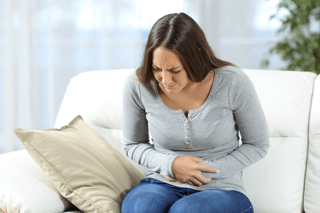 Woman sitting on sofa holding stomach in discomfort, stomach pain, digestive health issue