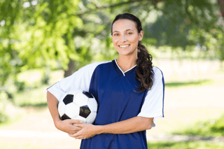 Smiling female soccer player in blue jersey holding soccer ball outdoors